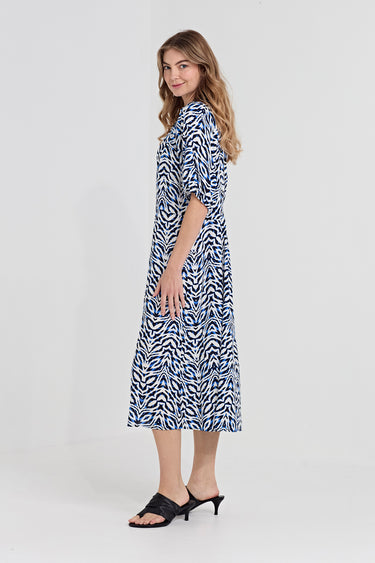A woman modeling a midi dress by Verge with a blue and white abstract wavy pattern, featuring elbow-length puff sleeves and a gathered waist, paired with black slide sandals against a light gray studio background.