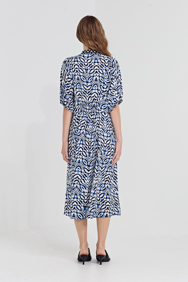 A woman modeling a midi dress by Verge with a blue and white abstract wavy pattern, featuring elbow-length puff sleeves and a gathered waist, paired with black slide sandals against a light gray studio background.