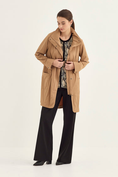 A woman modeling a camel-colored mid-length coat over a black and white patterned top and black wide-leg trousers by Verge, photographed against a plain light background.