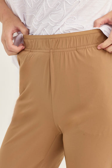Close-up of a person wearing tan or camel-colored pants by Verge, pulling the elastic waistband slightly outward with both hands. A white embroidered top is partially visible above the waistband.