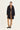 A model stands against a plain light background wearing a black mid-length coat over a tan sweater and tan pants by Verge, with black sneakers.