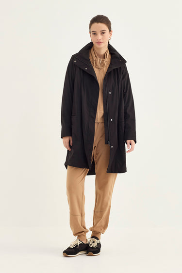 A model stands against a plain light background wearing a black mid-length coat over a tan sweater and tan pants by Verge, with black sneakers.