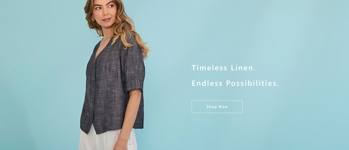 A woman wearing a dark gray linen short-sleeve button-up shirt and white pants by Verge, standing against a light blue background with text that reads 'Timeless Linen. Endless Possibilities.