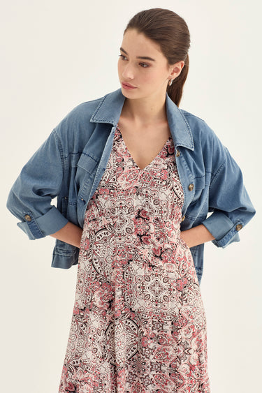 A woman wearing a light blue denim jacket over a sleeveless V-neck dress with a pink, beige, and black paisley pattern by Verge, posed against a plain off-white studio background.
