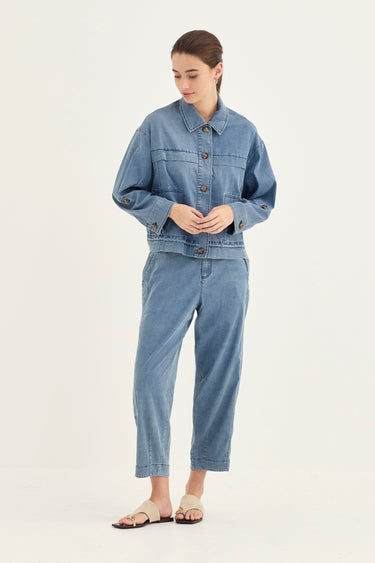 A woman modeling a light blue denim two-piece set consisting of a cropped button-up jacket and matching cropped pants by Verge, paired with beige flat sandals, against a plain off-white studio background.