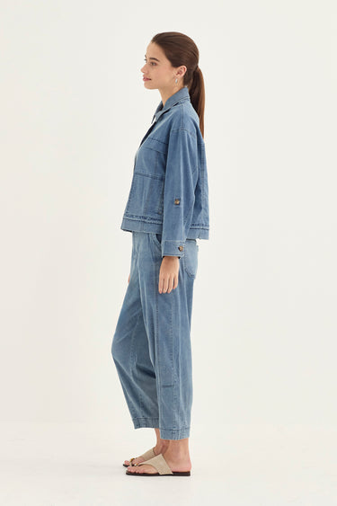 A woman stands in profile wearing a light blue denim jacket and matching wide-leg jeans, paired with beige flat sandals by Verge, against a plain off-white studio background.