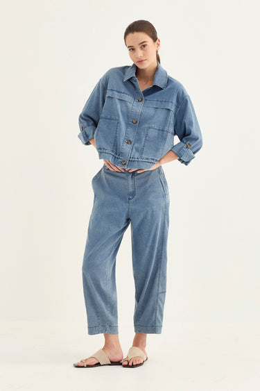 A woman modeling a light blue denim two-piece set consisting of a cropped button-up shirt jacket and matching cropped pants by Verge, paired with beige slide sandals, against a plain white studio background.