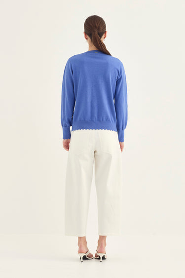 A woman viewed from the back wearing a periwinkle blue knit sweater with a scalloped hem, white cropped trousers by Verge, and black-and-white heeled sandals, against a plain off-white studio background.