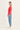 A woman stands in profile wearing a coral pink long-sleeve knit top, light blue jeans by Verge, and white sneakers against a plain off-white studio background.