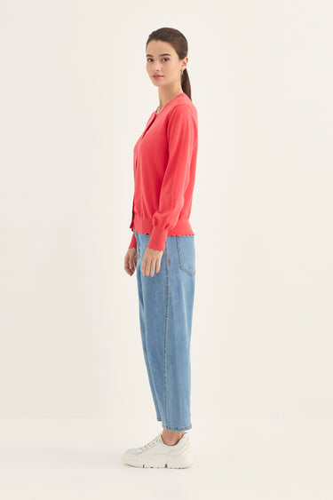 A woman stands in profile wearing a coral pink long-sleeve knit top, light blue jeans by Verge, and white sneakers against a plain off-white studio background.