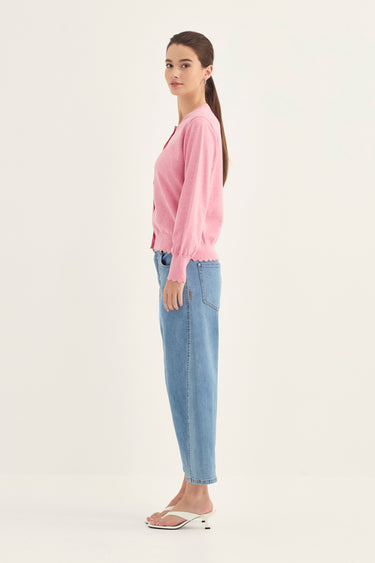 A woman stands in profile wearing a light pink long-sleeve knit top, light blue jeans by Verge, and white heeled sandals against a plain off-white studio background.
