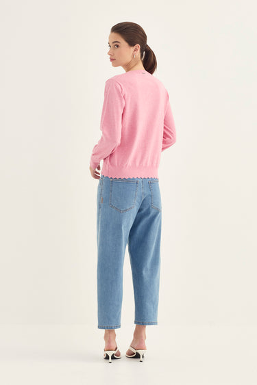 A woman is shown from the back wearing a light pink long-sleeve knit sweater with a scalloped hem and light blue cropped jeans by Verge, standing against a plain off-white studio background.