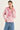 A woman modeling a pink and white striped long-sleeve top with a matching pink knit sweater by Verge draped over the shoulders, paired with blue jeans, against a plain light background.