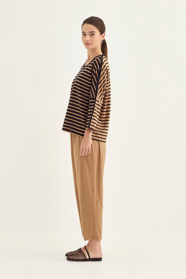 A woman stands in profile wearing a loose black and tan striped top with tan pants by Verge and flat woven sandals against a plain light background.