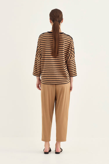 A woman viewed from the back wearing a brown and beige horizontally striped long-sleeve top and tan cropped pants by Verge, standing against a plain light background.