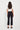 A woman shown from the back wearing black cropped trousers, a white short-sleeve top by Verge, and black flat sandals against a plain white studio background.