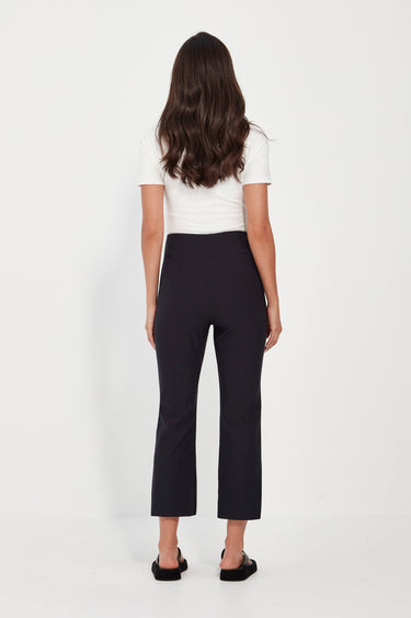 A woman shown from the back wearing black cropped trousers, a white short-sleeve top by Verge, and black flat sandals against a plain white studio background.