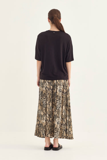 A woman viewed from the back wearing a black short-sleeve top and a beige and black snakeskin print midi skirt by Verge, paired with black flat sandals, against a plain off-white studio background.