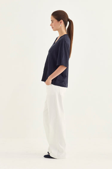 A woman in side profile wearing a dark navy blue short-sleeve top and white wide-leg pants by Verge, standing against a plain white studio background.