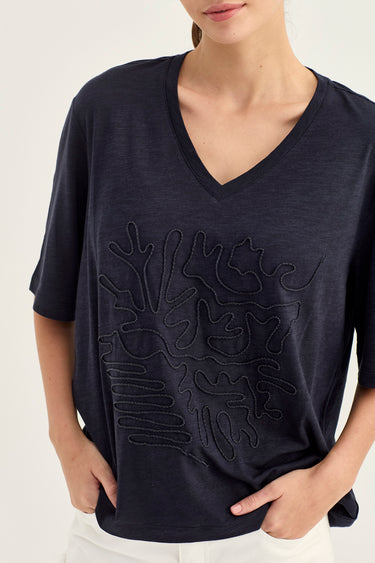 A woman wearing a dark navy blue V-neck short-sleeve top with an abstract tonal embroidered pattern on the front, paired with white pants by Vergeagainst a light off-white background.