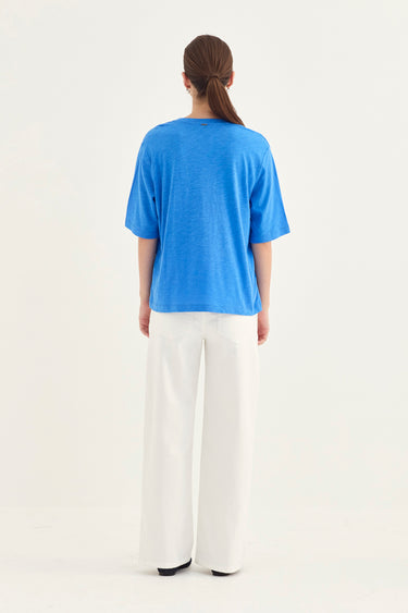 A woman viewed from behind wearing a bright blue short-sleeve t-shirt and white wide-leg trousers by Verge, standing against a plain white studio background.