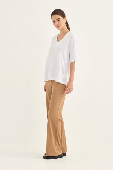 A woman modeling a white short-sleeve V-neck top and beige wide-leg pants by Verge against a plain off-white studio background.