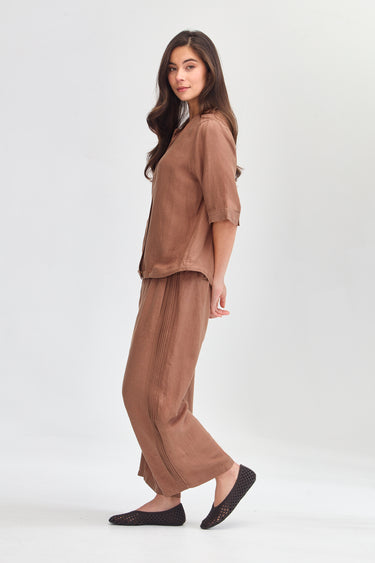 A woman modeling a matching two-piece lounge set in a warm taupe-brown color, featuring a short-sleeve top and wide-leg pants by Verge, paired with black slip-on flats against a light gray studio background.