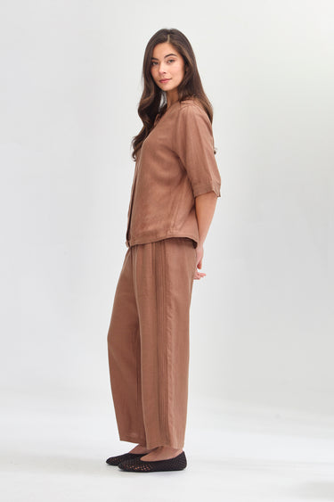 A woman modeling a matching two-piece lounge set in a warm brown color, featuring a short-sleeve top and wide-leg pants by Verge, photographed against a seamless light gray studio background.