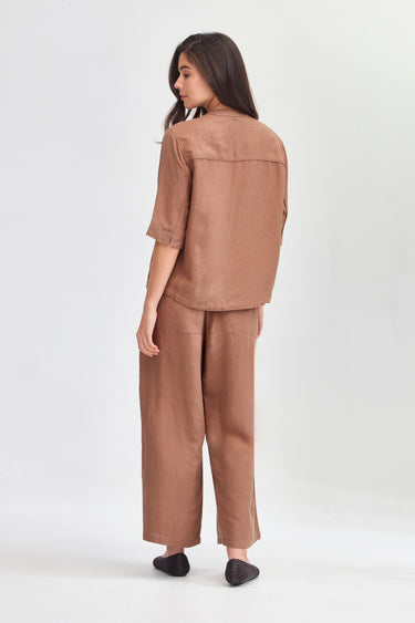 A woman is shown from the back wearing a matching two-piece linen set in a warm brownish-tan color, consisting of a short-sleeve button-up shirt and wide-leg trousers by Verge, against a plain light gray studio background.
