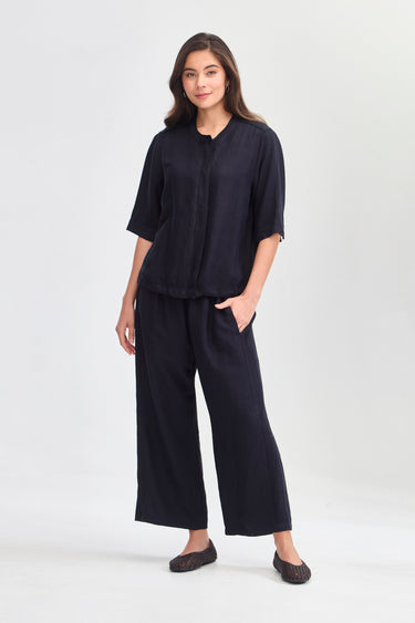 A woman modeling a matching dark navy two-piece linen set consisting of a short-sleeve button-up jacket by Verge and wide-leg trousers, paired with dark flat shoes, against a light gray studio background.