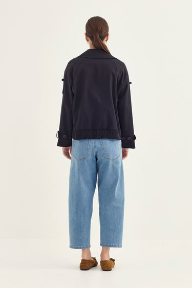 A model viewed from the back wearing a dark navy cropped jacket with sleeve tabs and light blue cropped jeans by Verge, paired with brown slip-on shoes, against a plain off-white studio background.