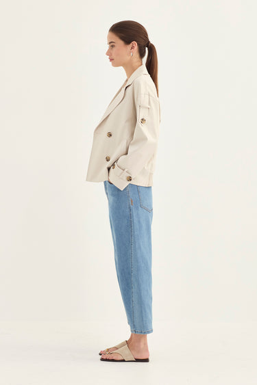 A woman in side profile wearing a light beige double-breasted blazer, light blue cropped jeans by Verge, and beige slide sandals, standing against a plain off-white studio background.