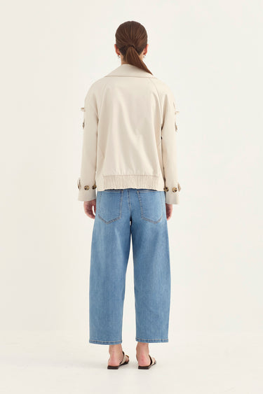 A woman viewed from behind wearing a cream-colored jacket with button details on the sleeves and light blue wide-leg cropped jeans by Verge, standing against a plain off-white studio background.