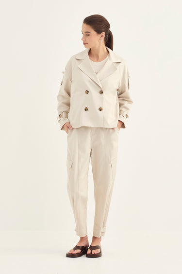 A woman modeling a light beige double-breasted jacket and matching trousers by Verge with dark brown sandals, photographed against a plain white studio background.