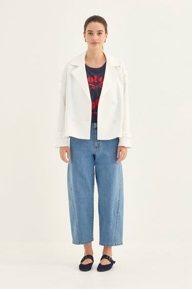 A model stands against a plain light background wearing a white double-breasted blazer over a dark graphic t-shirt, paired with wide-leg cropped blue jeans by Verge and black flat shoes by Verge.