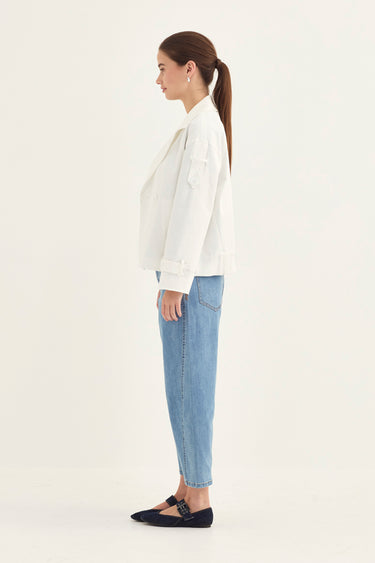 A woman in side profile wearing a white cropped jacket, light blue jeans by Verge, and black flat shoes against a plain off-white studio background.
