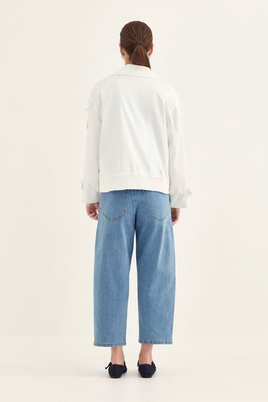 A model shown from the back wearing a white jacket, light blue cropped jeans by Verge, and black flats against a plain off-white studio background.