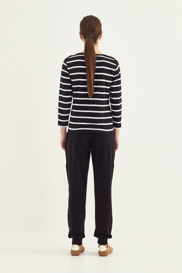 A woman viewed from behind wearing a black and white horizontally striped long-sleeve top and black pants by Verge, standing against a plain light background.