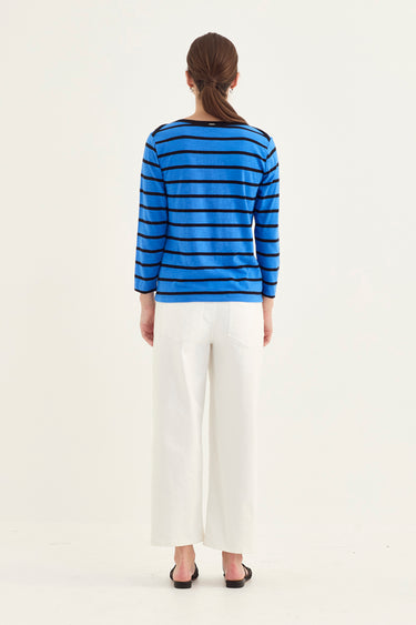 A woman viewed from the back wearing a blue and black striped long-sleeve top, white cropped wide-leg pants by Verge, and black sandals, against a plain light background.
