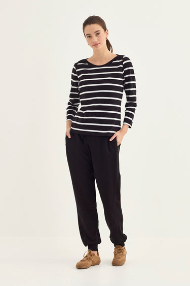 A woman modeling a black and white horizontally striped long-sleeve top with black jogger pants by Verge and brown shoes, standing against a plain white studio background.