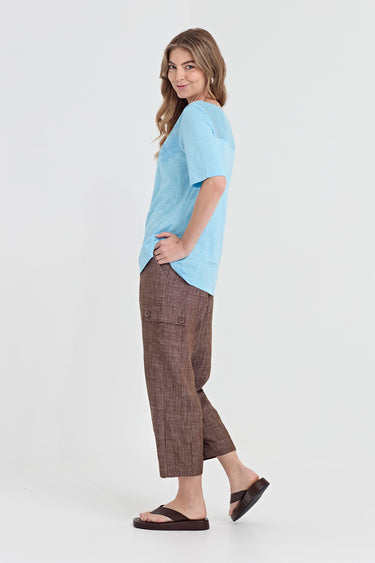 A woman modeling a light blue short-sleeve t-shirt and brown cropped plaid pants by Verge with dark brown sandals, standing against a plain white studio background.