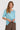 A woman modeling a light blue short-sleeve top with a sheer floral lace overlay on the chest, paired with brown textured pants by Verge, against a solid light blue background.