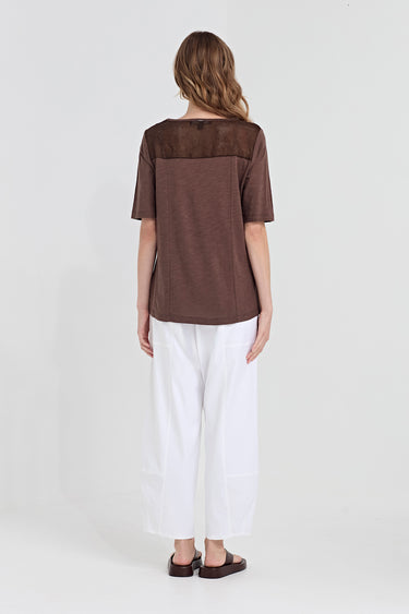 A woman viewed from the back wearing a brown short-sleeve top with a wide shoulder yoke and white wide-leg cropped pants by Verge, standing against a plain light gray studio background.