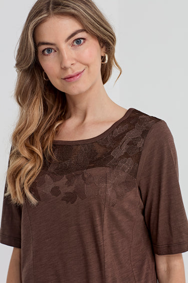 A woman wearing a brown short-sleeve top by Verge with a sheer lace yoke across the chest, photographed against a light gray studio background.