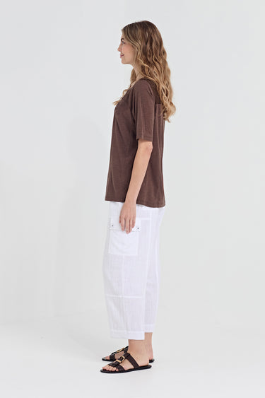 A woman shown in profile wearing a brown short-sleeve top, white wide-leg pants by Verge, and black sandals against a plain light gray studio background.
