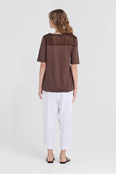 A woman viewed from the back wearing a brown short-sleeve top with a sheer mesh yoke and white cropped pants by Verge, standing against a plain light gray studio background.