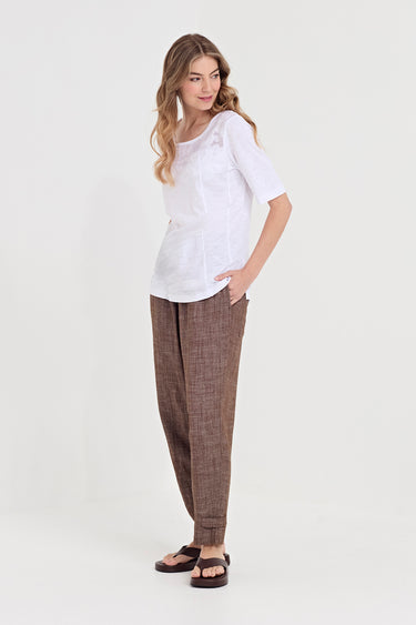 A woman modeling a white short-sleeve top and brown textured trousers by Verge with dark brown sandals, standing against a plain light gray studio background.