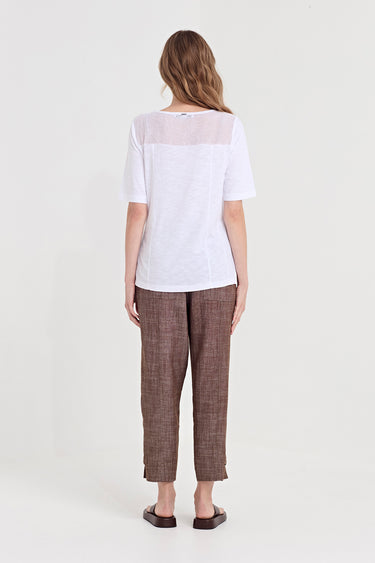 A woman viewed from the back wearing a white short-sleeve top with a sheer yoke and brown textured cropped trousers by Verge, standing against a plain light gray studio background.