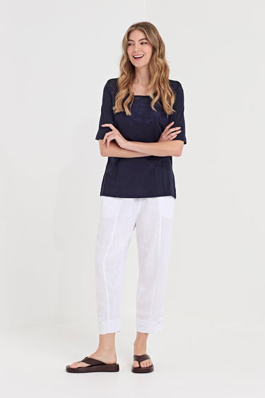 A woman modeling a dark navy blue short-sleeve top and white cropped pants by Verge with brown sandals, standing against a plain light gray studio background.