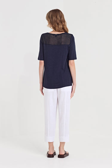 A woman viewed from the back wearing a navy blue short-sleeve top with a sheer yoke and white cropped pants by Verge, standing against a plain light gray studio background.